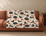 Pumpkin Patch Cats Halloween blanket on a rust-colored sofa, with charcoal black cats in three poses and orange pumpkins on a warm cream background.