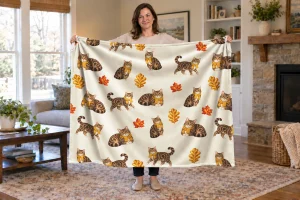 Woman holding an open Autumn Cat Naps blanket with tabby cats in mustard scarves and autumn leaves.