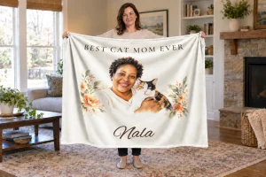 Woman holding a Best Cat Mom Ever blanket with a watercolor portrait of Nala, her calico cat, and herself, with Nala’s name below.