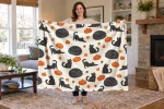 Woman holding an open Pumpkin Patch Cats blanket with black cats and orange pumpkins on a cream background.