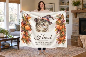 Woman holding Hazel’s cream Halloween cat blanket in an autumn living room, showing her witch-hat portrait, name, and Autumn Looks Good on You.