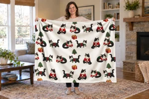 Woman holding an open Sweater Weather Cats blanket with tuxedo cats in red and green sweaters, fir trees, and gifts.