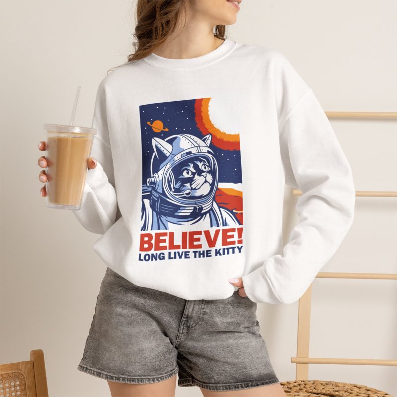 Person wearing a crewneck sweatshirt showing a gray tabby astronaut, a ringed golden planet and stars on deep blue