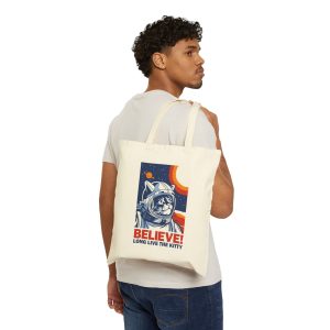 Person carrying a natural canvas tote showing a gray tabby astronaut, a ringed golden planet and stars on deep blue
