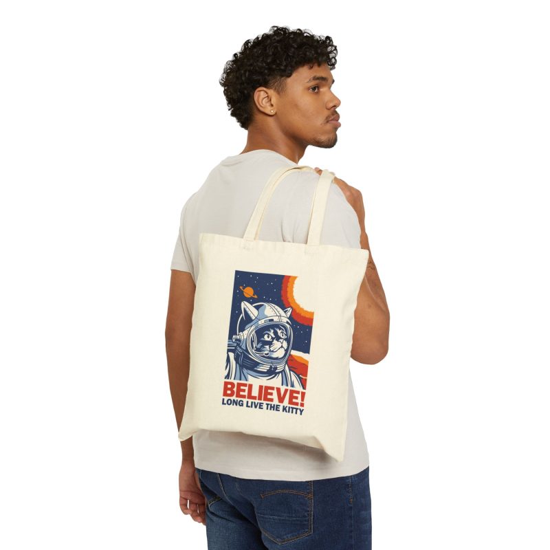 Person carrying a natural canvas tote showing a gray tabby astronaut, a ringed golden planet and stars on deep blue