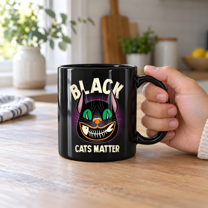 Person holding a glossy black ceramic mug showing a grinning black cat with huge green eyes, orange mouth outlines and a purple halo