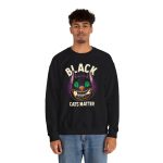 Person wearing a crewneck sweatshirt showing a grinning black cat with huge green eyes, orange mouth outlines and a purple halo