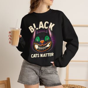 Person wearing a crewneck sweatshirt showing a grinning black cat with huge green eyes, orange mouth outlines and a purple halo