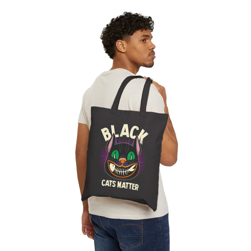 Person carrying a black canvas tote showing a grinning black cat with huge green eyes, orange mouth outlines and a purple halo