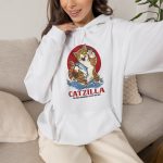 Person wearing a hoodie showing a giant brown tabby holding a blue fish above waves in front of a red sun disc