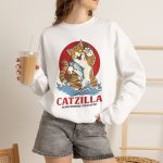 Person wearing a crewneck sweatshirt showing a giant brown tabby holding a blue fish above waves in front of a red sun disc