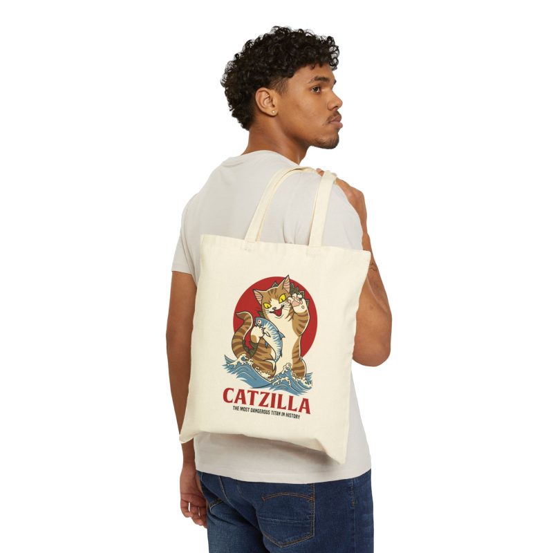 Person carrying a natural canvas tote showing a giant brown tabby holding a blue fish above waves in front of a red sun disc