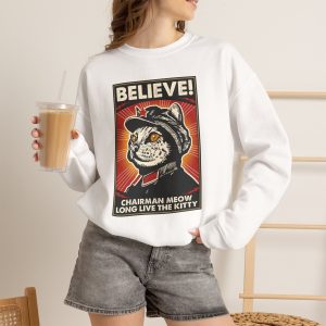 Person wearing a crewneck sweatshirt showing a stern black-and-cream cat in a red-star cap with orange-red sunburst rays and a black frame