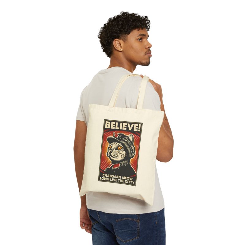 Person carrying a natural canvas tote showing a stern black-and-cream cat in a red-star cap with orange-red sunburst rays and a black frame