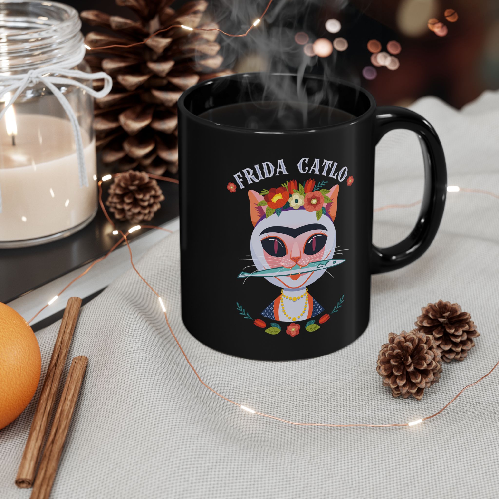 Glossy black ceramic mug showing a pale pink cat with a unibrow, a flower crown, gold beads and a teal fish