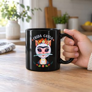 Person holding a glossy black ceramic mug showing a pale pink cat with a unibrow, a flower crown, gold beads and a teal fish