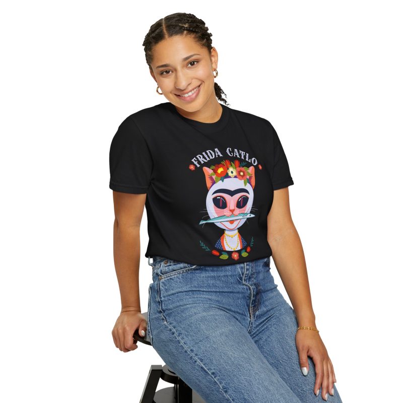 Person wearing a tee with a pale pink cat with a unibrow, a flower crown, gold beads and a teal fish printed on the front