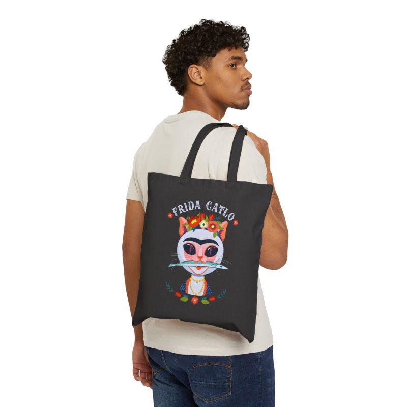 Person carrying a black canvas tote with a pale pink cat with a unibrow, a flower crown, gold beads and a teal fish facing outward