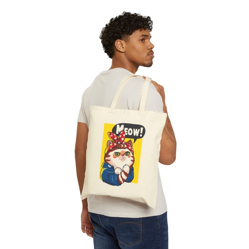 Person carrying a natural canvas tote with a white and ginger cat flexing its paw in a blue work shirt and red polka-dot bandana on a yellow panel facing outward