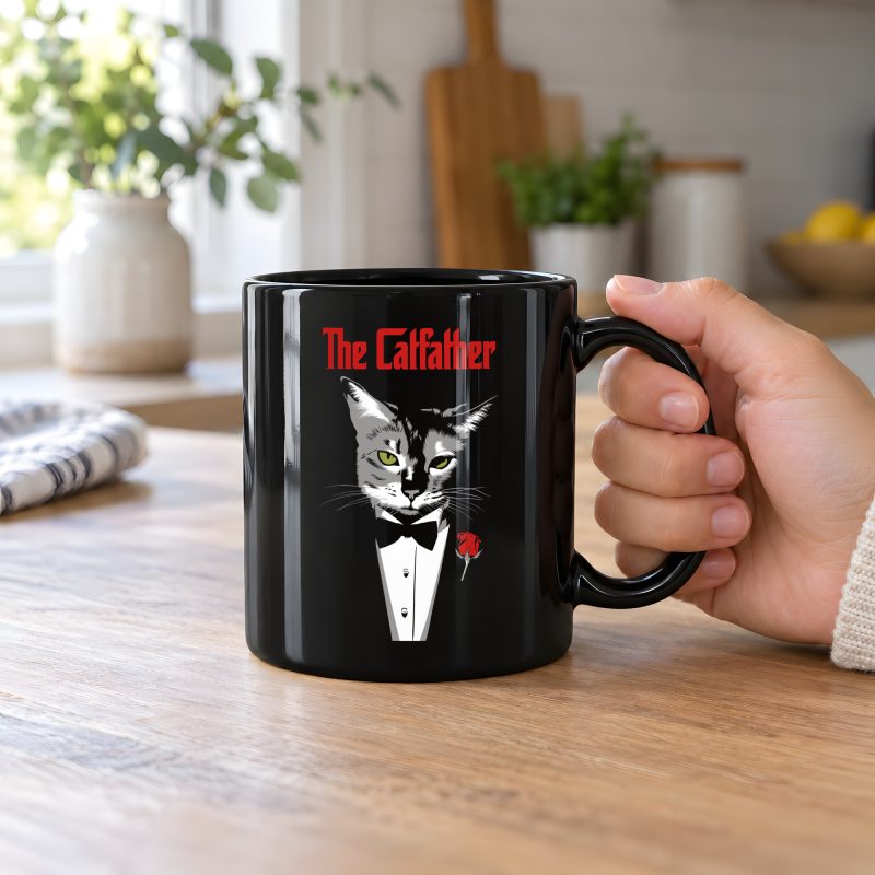 Person holding a glossy black ceramic mug showing a gray cat with green eyes in a white tuxedo shirt and black bow tie with a red rose