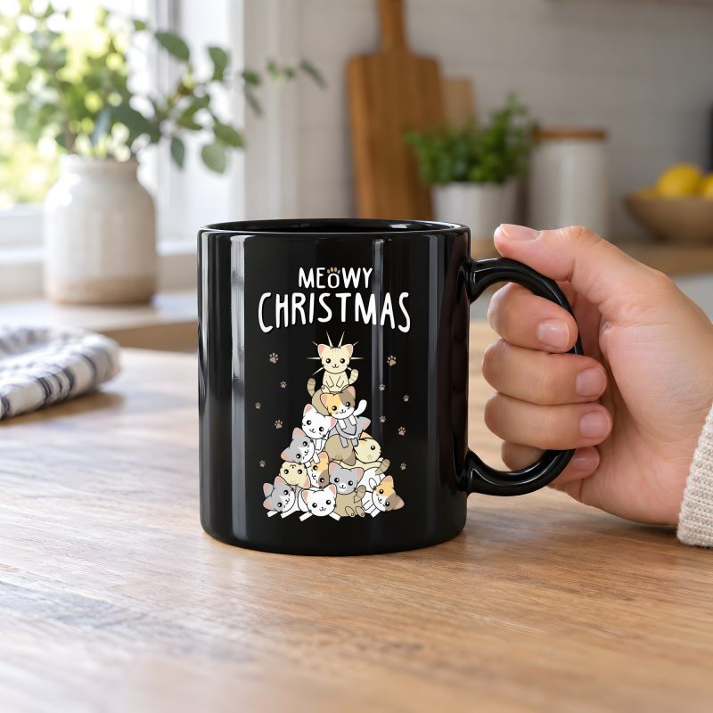 Person holding a glossy black ceramic mug with white, gray, cream and ginger kittens stacked into a Christmas tree, surrounded by paw prints beneath white lettering