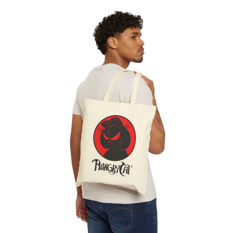 Person carrying a natural canvas tote with a sly black cat silhouette inside a red circle facing outward