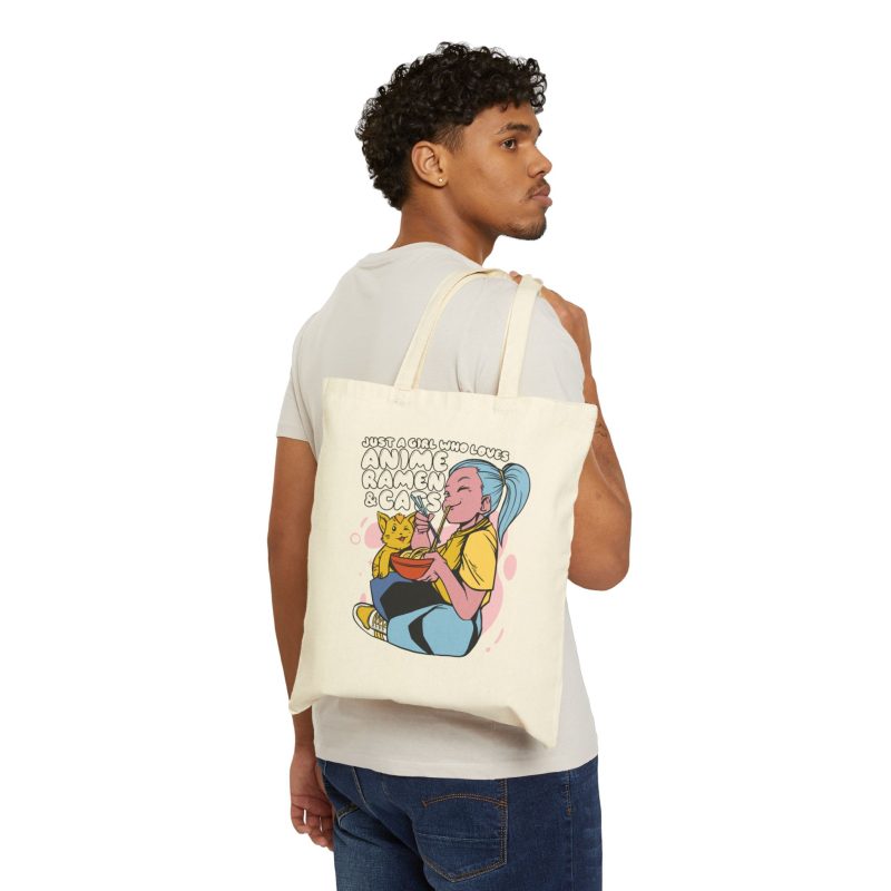 Person carrying a natural canvas tote showing a blue-haired girl eating ramen beside a golden ginger cat