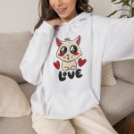 Person wearing a hoodie with a cream cat with glossy black eyes and red cheeks between two red hearts above black lettering printed on the front