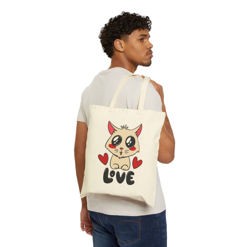 Person carrying a natural canvas tote with a cream cat with glossy black eyes and red cheeks between two red hearts above black lettering facing outward