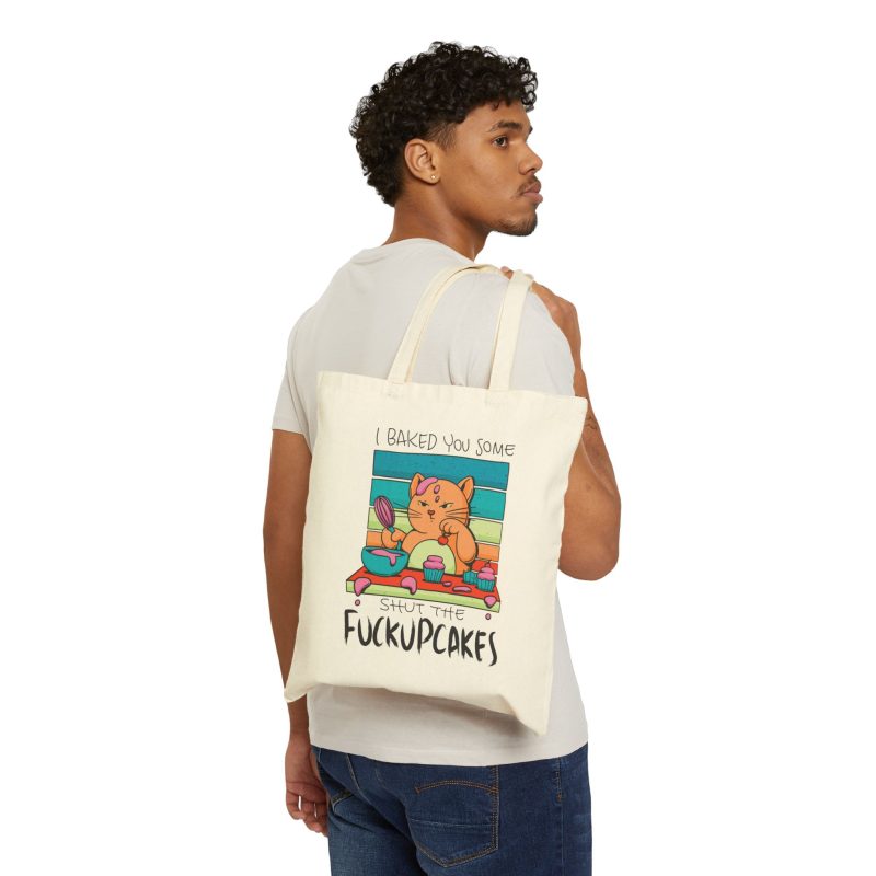 Person carrying a natural canvas tote showing a ginger baker cat with a pink whisk, cupcakes and turquoise retro stripes