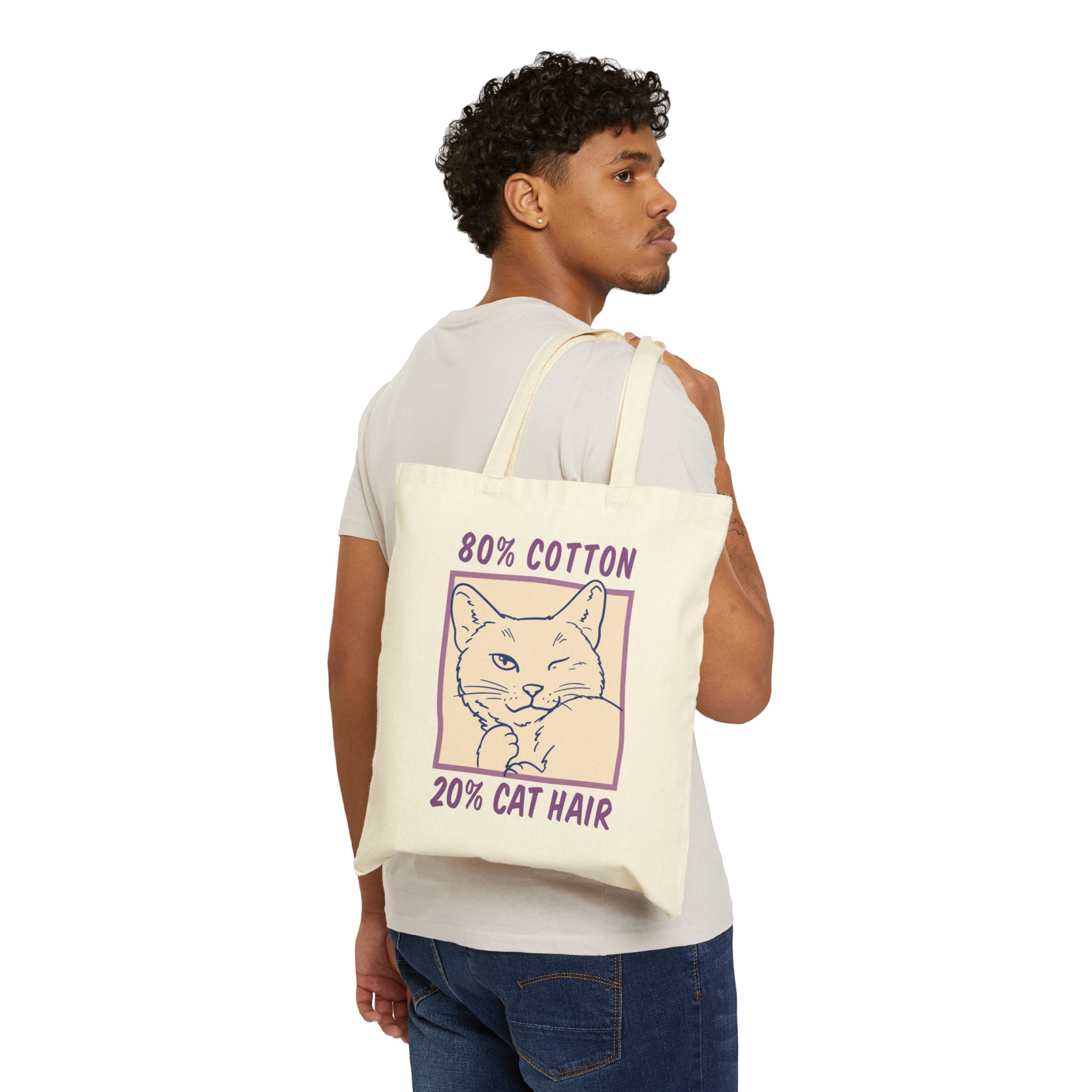 Person carrying a natural canvas tote showing a winking white cat drawn in blue inside a cream square with purple lettering