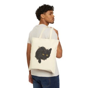 Person carrying a natural canvas tote showing an illustrated black cat with yellow-green eyes and a paw reaching through a gray torn-hole effect