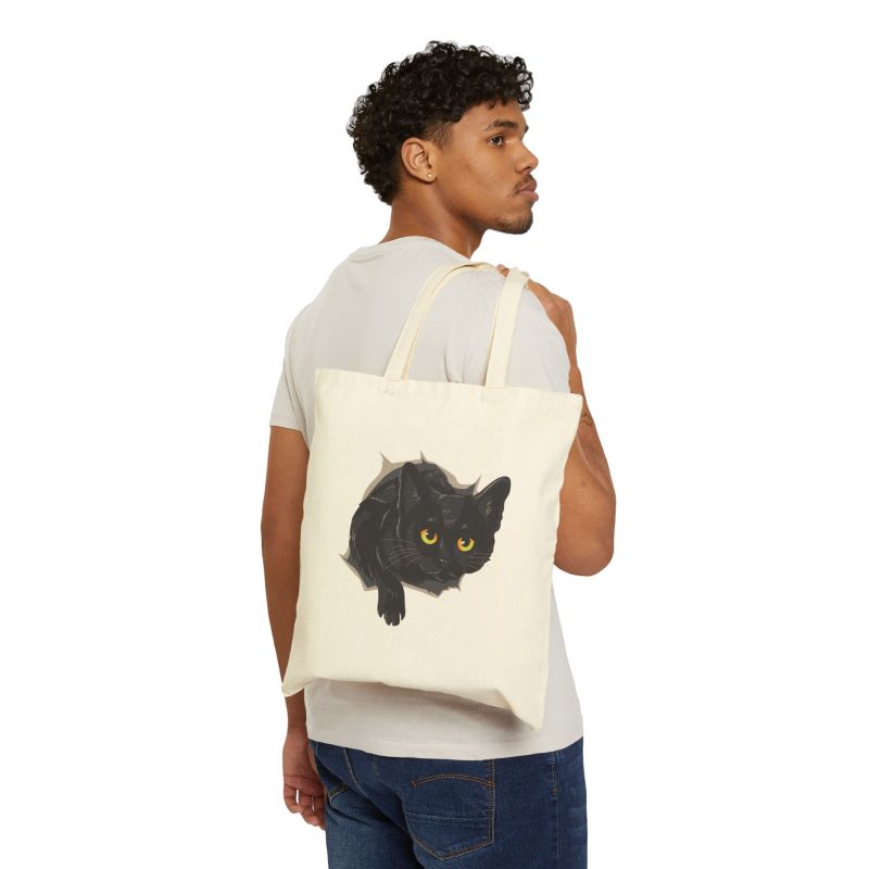 Person carrying a natural canvas tote showing an illustrated black cat with yellow-green eyes and a paw reaching through a gray torn-hole effect