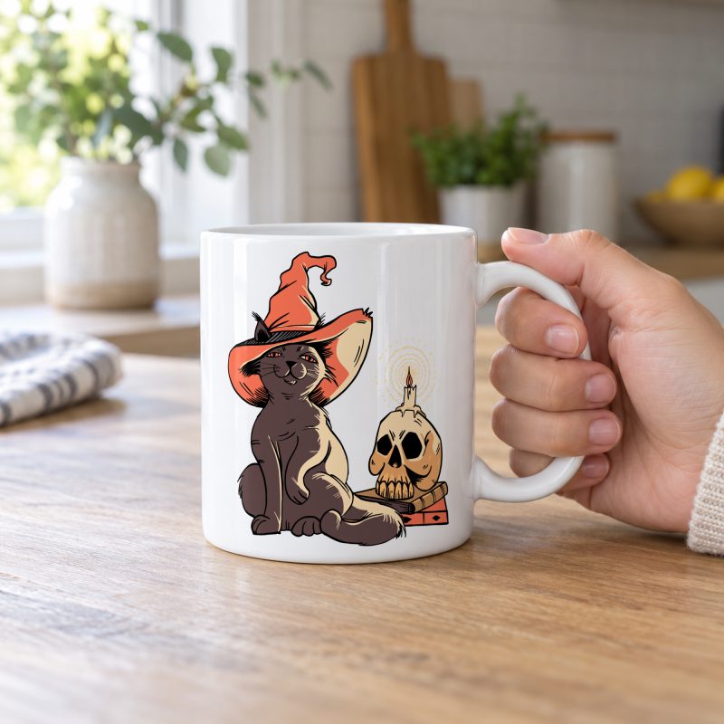 Person holding a glossy white ceramic mug showing a brown cat in an orange witch hat beside a skull, lit candle and stacked spell books