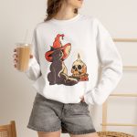 Person wearing a crewneck sweatshirt with a brown cat in an orange witch hat beside a skull, lit candle and stacked spell books printed on the front