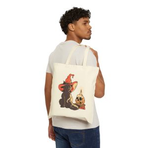 Person carrying a natural canvas tote showing a brown cat in an orange witch hat beside a skull, lit candle and stacked spell books