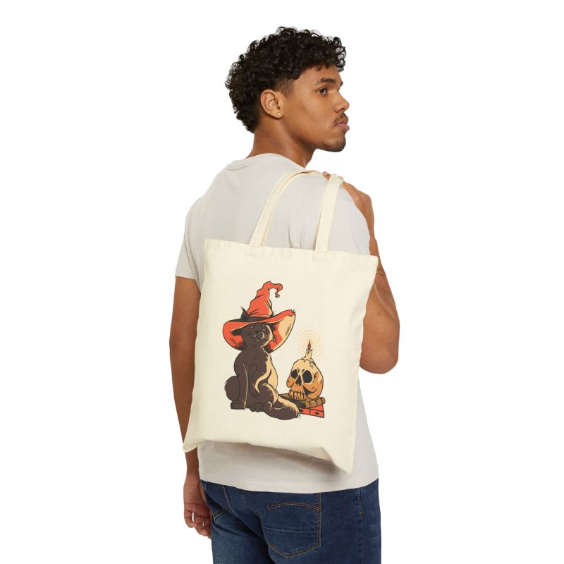 Person carrying a natural canvas tote showing a brown cat in an orange witch hat beside a skull, lit candle and stacked spell books
