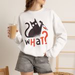 Person wearing a crewneck sweatshirt showing a wide-eyed black cat with a kitchen knife and black and orange lettering on the front Person wearing a crewneck sweatshirt showing a wide-eyed black cat with a kitchen knife and black and orange lettering on the front