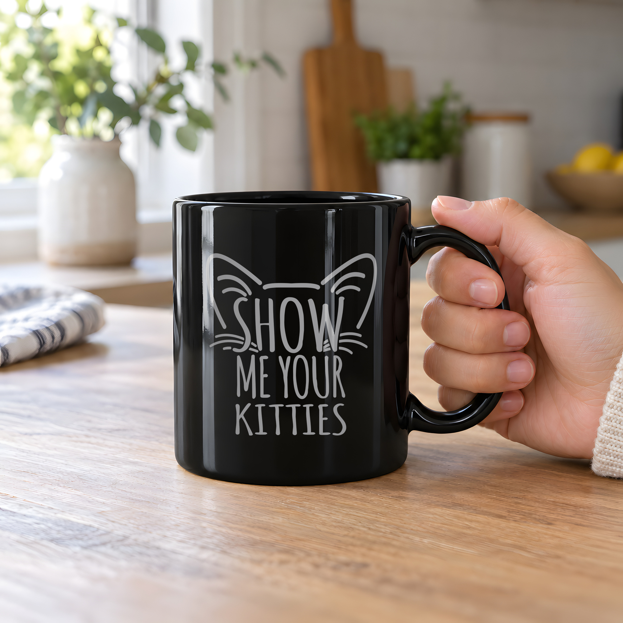 Person holding a glossy black ceramic mug showing gray cat-ear and whisker lettering
