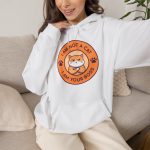 Person wearing a hoodie with an unimpressed ginger tabby with crossed arms inside an orange badge with dark purple lettering and two paw prints printed on the front Person wearing a hoodie with an unimpressed ginger tabby with crossed arms inside an orange badge with dark purple lettering and two paw prints printed on the front