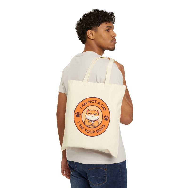 Person carrying a natural canvas tote with an unimpressed ginger tabby with crossed arms inside an orange badge with dark purple lettering and two paw prints facing outward