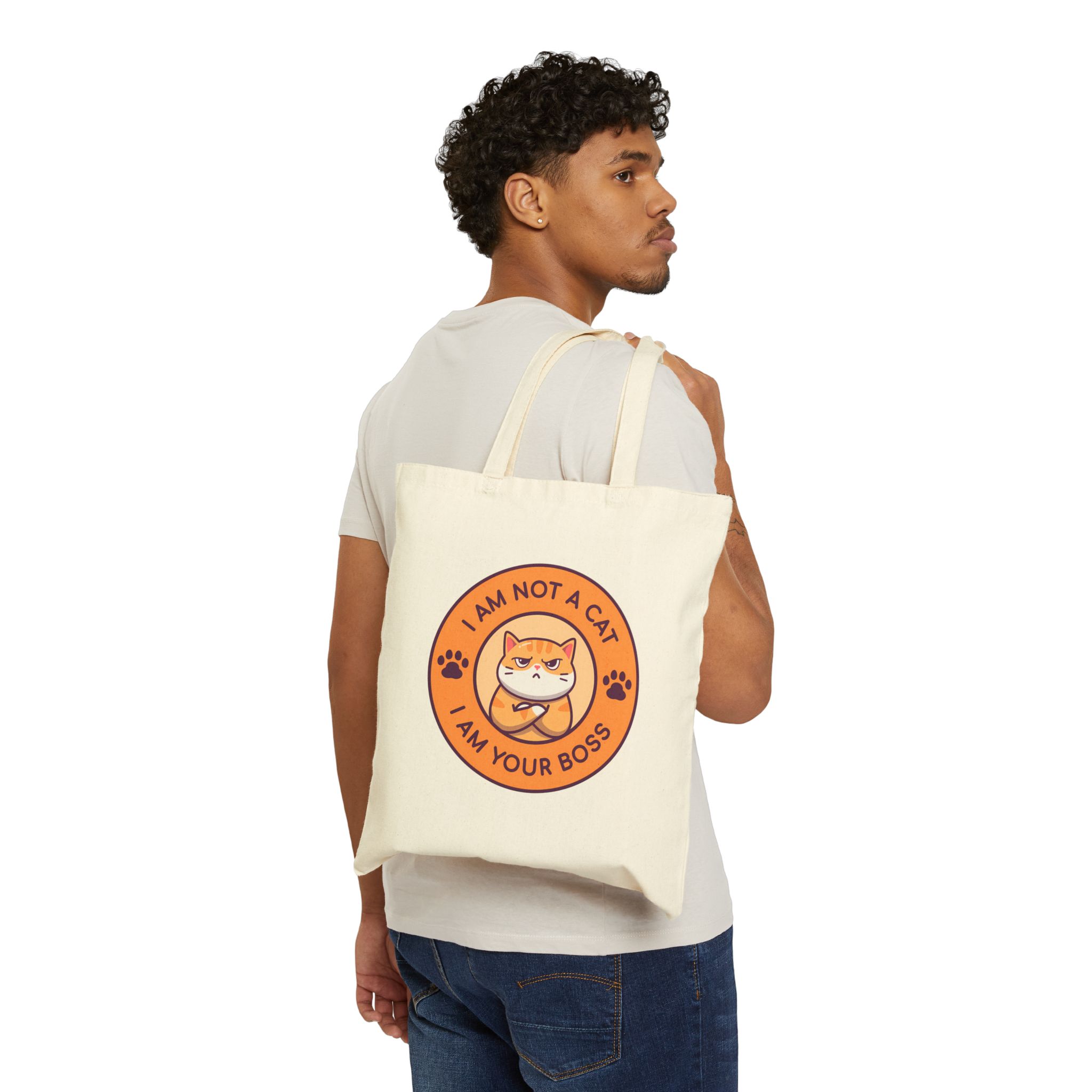 Person carrying a natural canvas tote with an unimpressed ginger tabby with crossed arms inside an orange badge with dark purple lettering and two paw prints facing outward
