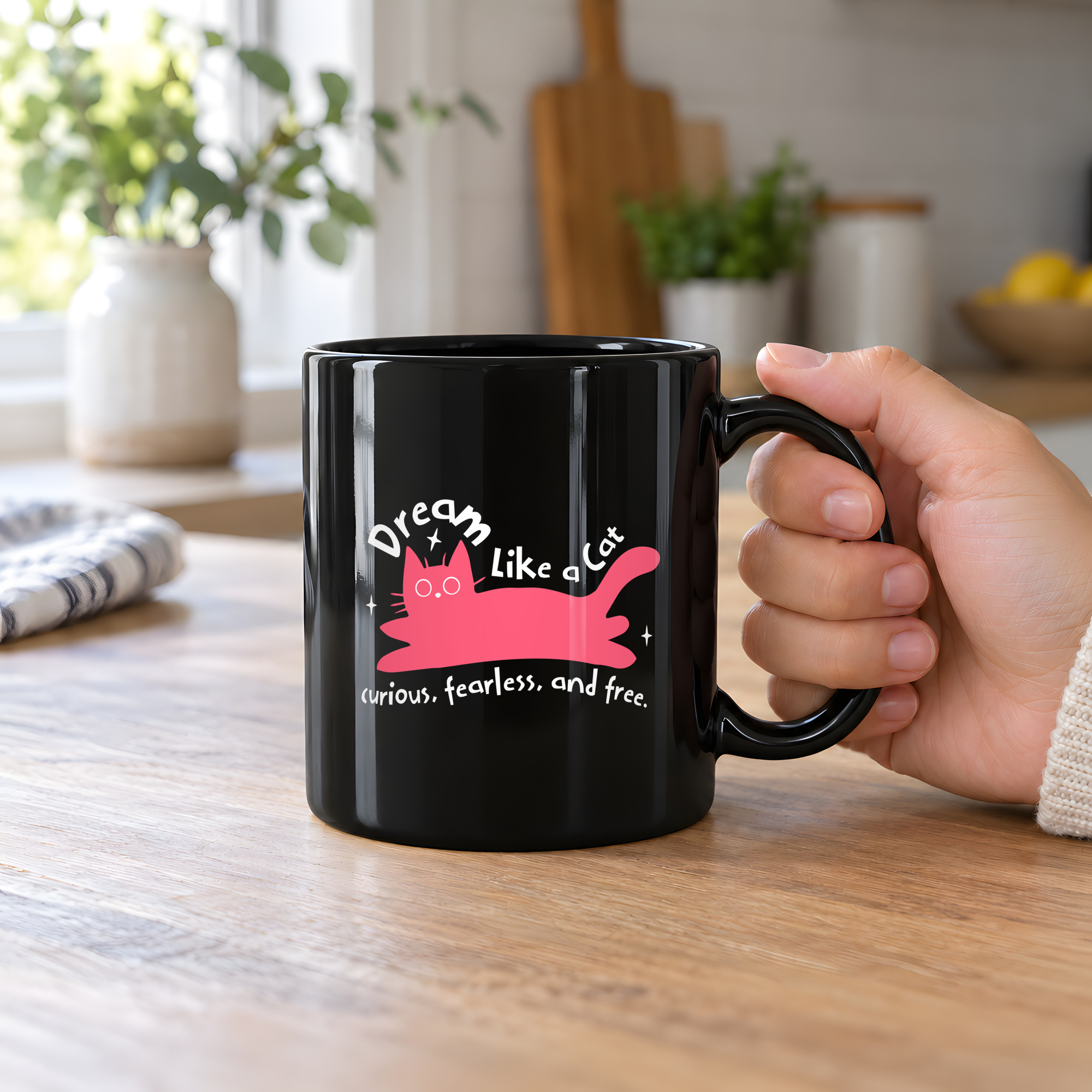 Person holding a glossy black ceramic mug showing a pink reclining cat with a raised tail, round eyes and white sparkles