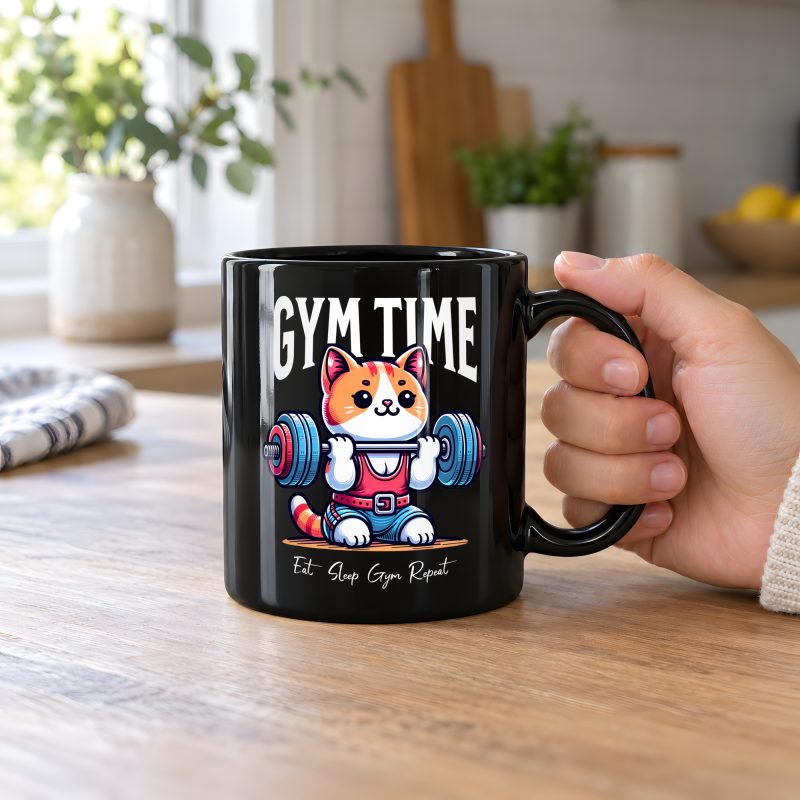 Person holding a glossy black ceramic mug showing an orange-and-white kitten lifting a barbell with blue and pink weight plates