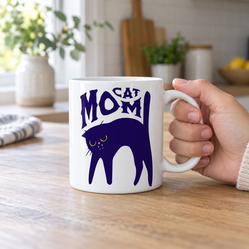 Person holding a white ceramic mug with a navy arched cat and wavy lettering graphic