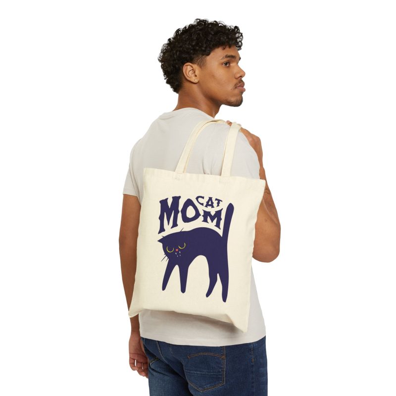 Person carrying a natural canvas tote with the navy arched cat and wavy lettering graphic facing outward