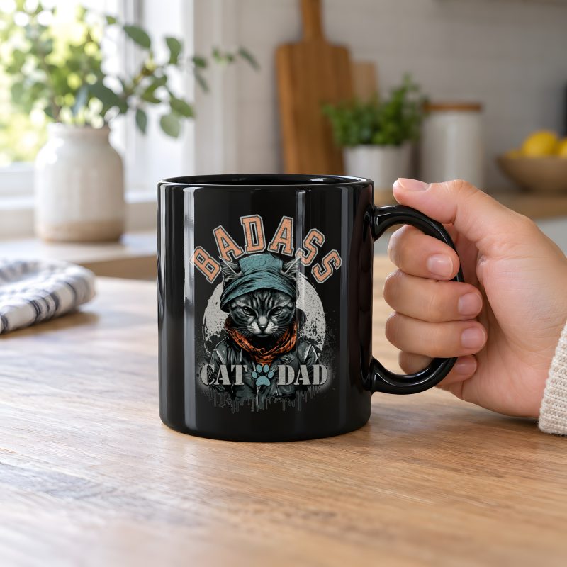 Person holding a glossy black ceramic mug with a tabby in a cap, jacket and scarf graphic