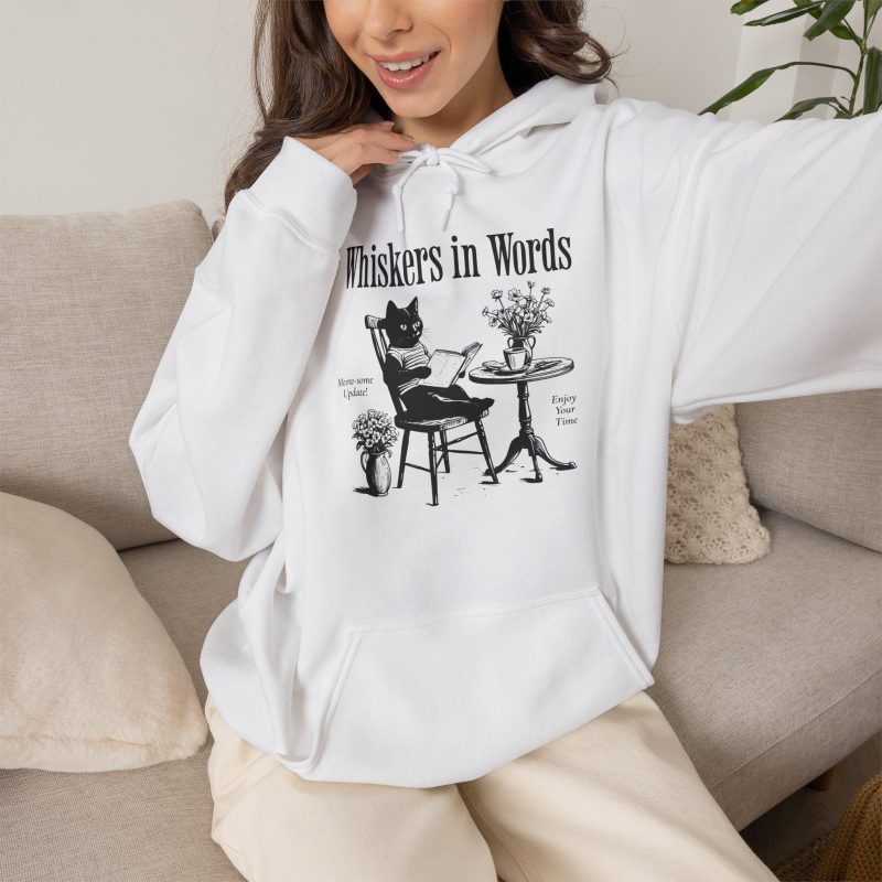 Person wearing a hoodie with a cat reading a book at a café table graphic on the front