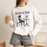 Person wearing a crewneck sweatshirt with a cat reading a book at a café table graphic on the front Person wearing a crewneck sweatshirt with a cat reading a book at a café table graphic on the front