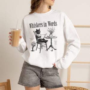 Person wearing a crewneck sweatshirt with a cat reading a book at a café table graphic on the front